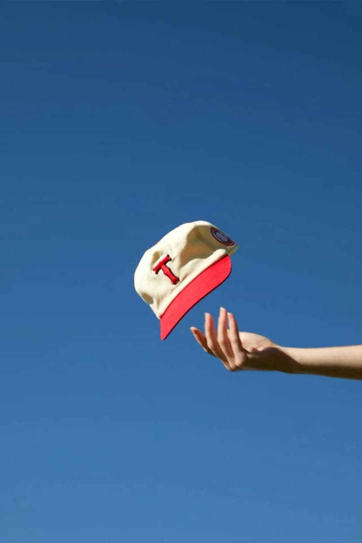 Minimalist product photo of a cream baseball cap with a red brim and red embroidered “T,” floating above an outstretched hand against a vast cloudless blue sky. Bright midday sunlight with crisp shadows, clean negative space, airy summery mood, sharp focus.