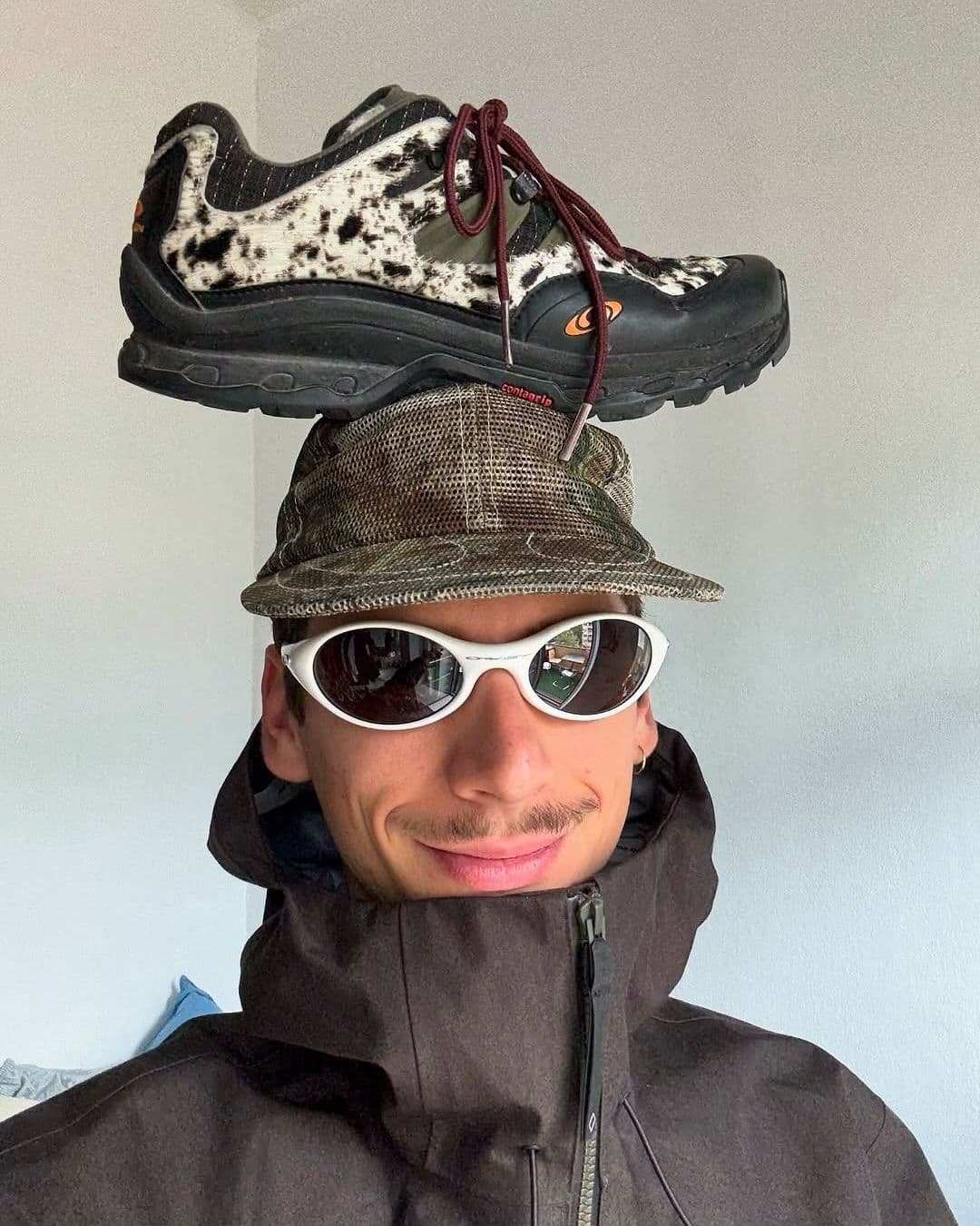 Surreal product photo of a rugged trail sneaker with a black rubber sole, white-and-black speckled upper and burgundy laces, balanced atop a camo bucket hat like a pedestal. Shot indoors against a plain light-gray wall with soft natural daylight, crisp detail and a playful, offbeat streetwear mood.
